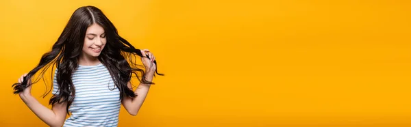 Brunette femme durcissant bouclés cheveux longs et souriant isolé sur jaune, panoramique shot — Photo de stock