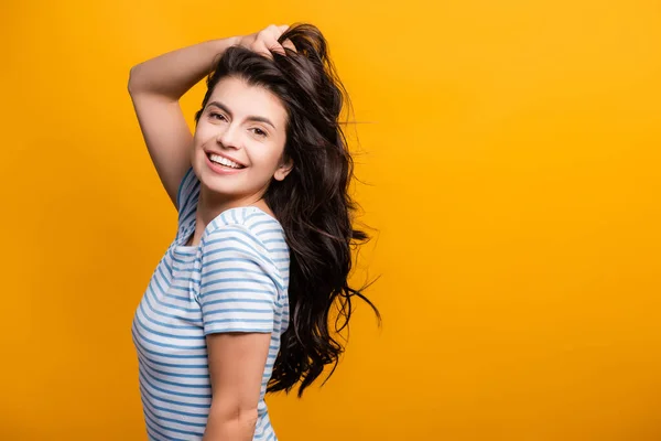 Vista lateral de la mujer morena endureciendo el pelo largo rizado y sonriendo aislado en amarillo - foto de stock