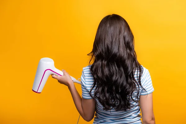 Vue arrière de la femme brune avec des boucles tenant sèche-cheveux isolé sur jaune — Photo de stock