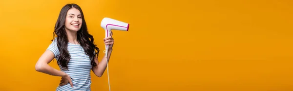 Brunette woman with curls holding hairdryer isolated on yellow, panoramic shot — Stock Photo