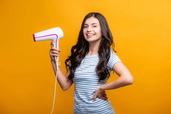 Brunette woman with curls holding hairdryer isolated on yellow — Stock Photo