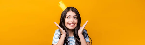 Brunette woman in paper crown smiling and looking away isolated on yellow, panoramic shot — Stock Photo
