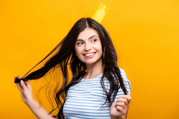 Morena mujer en papel corona tocando el pelo y sonriendo aislado en amarillo - foto de stock
