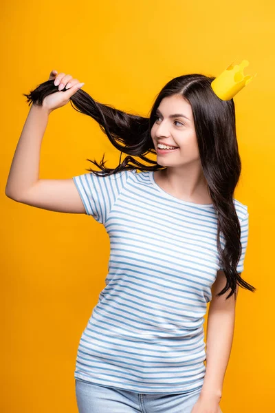 Morena mujer en papel corona sosteniendo el pelo y sonriendo aislado en amarillo - foto de stock
