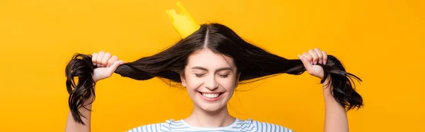 Morena mujer en corona de papel con los ojos cerrados sosteniendo el pelo y sonriendo aislado en amarillo, tiro panorámico - foto de stock