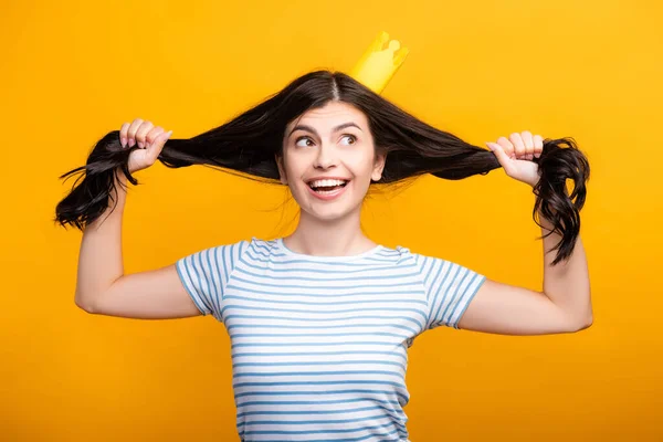 Morena mujer en papel corona sosteniendo el pelo y sonriendo aislado en amarillo - foto de stock