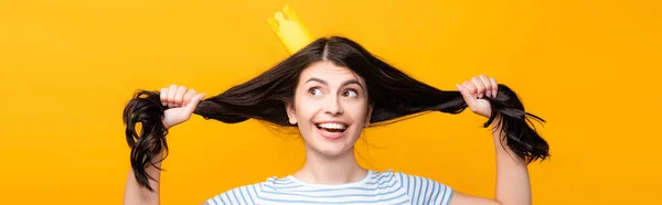 Brunette femme en papier couronne tenant les cheveux et souriant isolé sur jaune, panoramique shot — Photo de stock