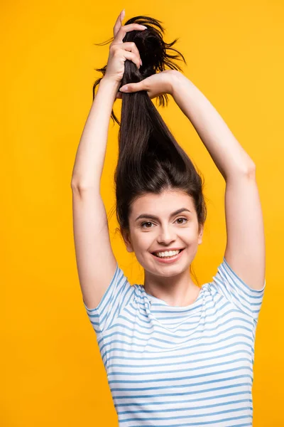 Morena mujer sosteniendo el pelo por encima de la cabeza y sonriendo aislado en amarillo - foto de stock