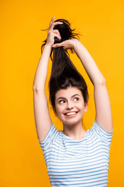 Brunette woman holding hair above head and smiling isolated on yellow — Stock Photo