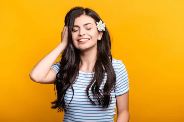 Brunette jeune femme avec des fleurs dans les cheveux et les yeux fermés souriant isolé sur jaune — Photo de stock