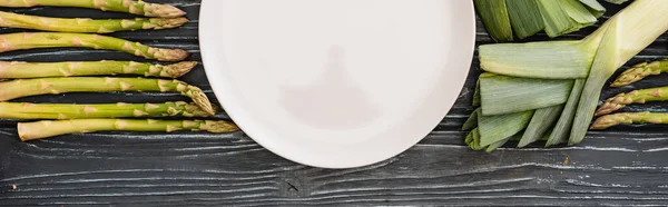 Top view of fresh green leek, asparagus and empty plate on wooden surface, panoramic shot — Stock Photo