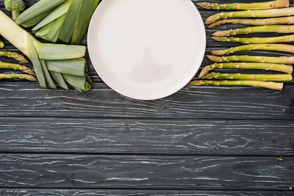 Vista dall'alto di porro verde fresco, asparagi e piatto vuoto su superficie in legno — Foto stock