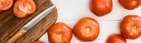 Vista superior de tomates maduros sobre tabla de cortar con cuchillo sobre mesa de madera blanca, plano panorámico - foto de stock