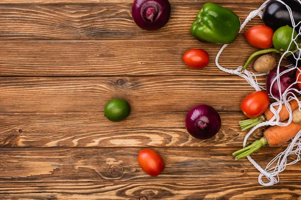 Top view of fresh ripe vegetables scattered from string bag on wooden table — Stock Photo