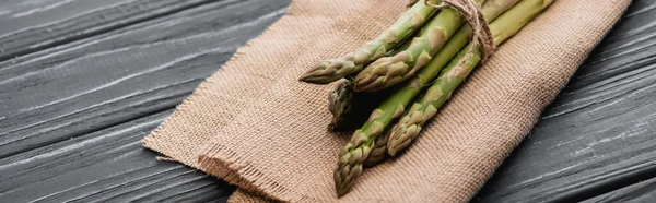 Bundle of fresh green asparagus on burlap on wooden surface, panoramic shot — Stock Photo