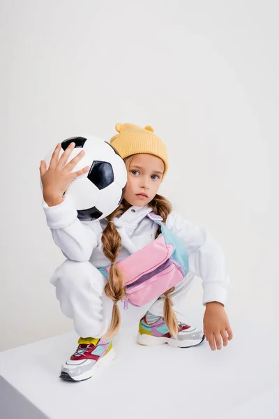 Rubia chica en ropa deportiva posando con pelota de fútbol en cubo aislado en blanco - foto de stock