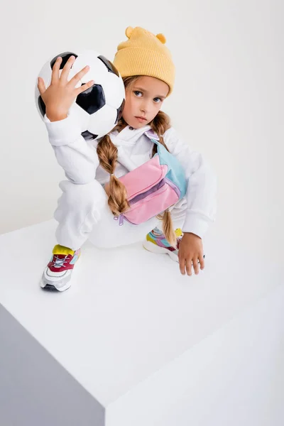 Rubia chica en ropa deportiva posando con pelota de fútbol en cubo aislado en blanco - foto de stock