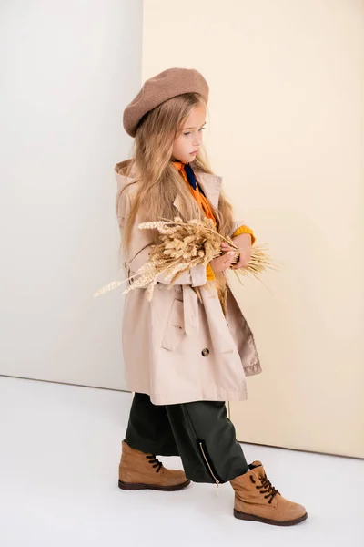 Vue latérale de la fille blonde à la mode en tenue d'automne marchant avec des pointes de blé sur fond beige et blanc — Photo de stock