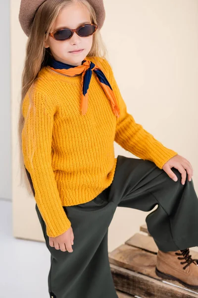 Fille blonde à la mode en tenue d'automne et lunettes de soleil posant sur une boîte en bois sur fond beige et blanc — Photo de stock