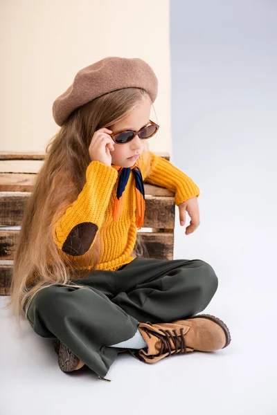 Chica rubia de moda en traje de otoño y gafas de sol posando cerca de caja de madera sobre fondo beige y blanco - foto de stock