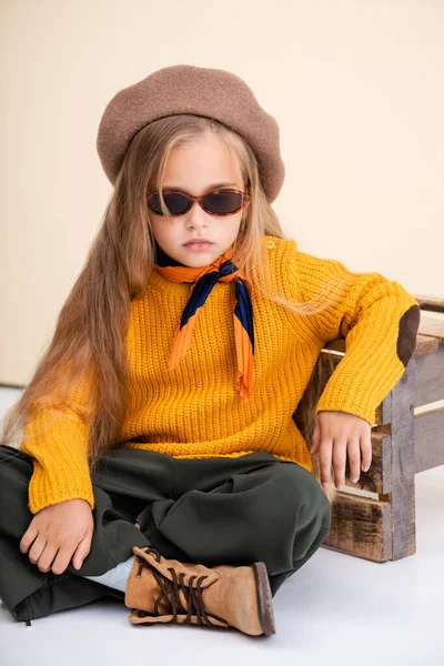 Moda ragazza bionda in abito autunno e occhiali da sole posa vicino scatola di legno su sfondo beige e bianco — Foto stock