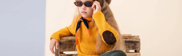 Modisches blondes Mädchen im Herbst-Outfit und Sonnenbrille posiert in der Nähe einer Holzkiste auf beige-weißem Hintergrund, Panoramaaufnahme — Stockfoto