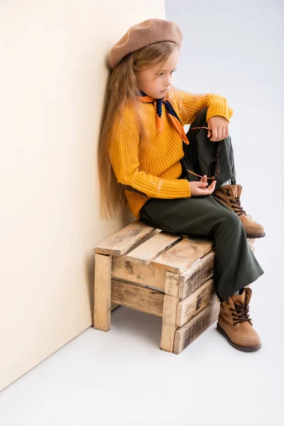 Fashionable blonde girl in autumn outfit with sunglasses sitting on wooden box on beige and white background — Stock Photo