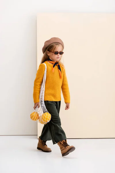 Fashionable blonde girl in autumn outfit and sunglasses walking with grapefruits in string bag on beige and white background — Stock Photo