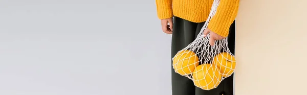 Vue recadrée de fille à la mode en tenue d'automne posant avec des pamplemousses dans un sac à ficelle sur fond beige et blanc, prise de vue panoramique — Photo de stock