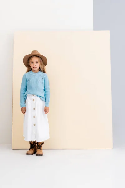 Ragazza bionda alla moda in cappello marrone e stivali, gonna bianca e maglione blu vicino alla parete beige — Foto stock