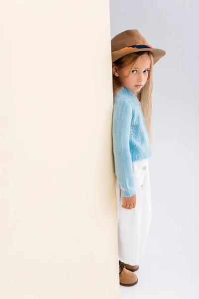Vista lateral de la chica rubia de moda en sombrero marrón y botas, falda blanca y suéter azul cerca de la pared beige - foto de stock