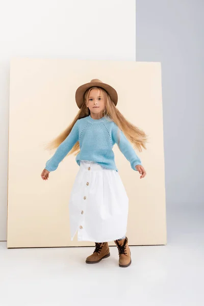 Fashionable blonde girl in brown hat and boots, white skirt and blue sweater spinning near beige wall — Stock Photo