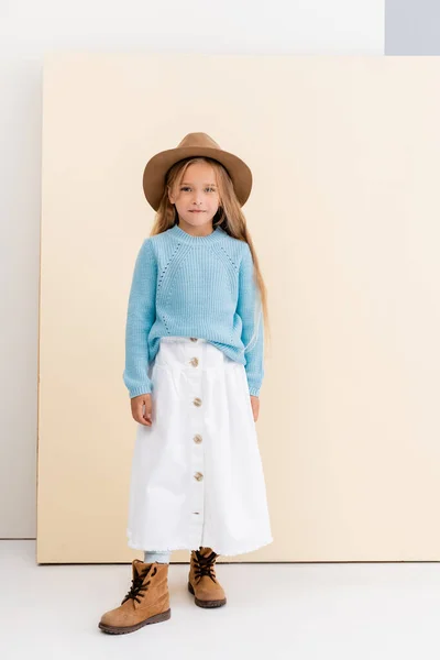 Fille blonde à la mode en chapeau et bottes marron, jupe blanche et pull bleu près du mur beige — Photo de stock