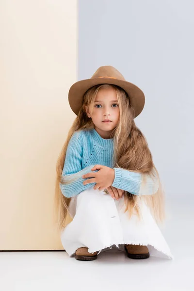 Fashionable blonde girl in brown hat and boots, white skirt and blue sweater sitting near beige wall — Stock Photo
