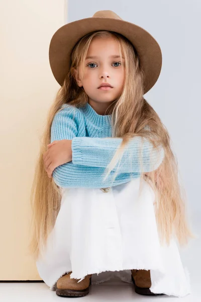 Fille blonde à la mode en chapeau brun et bottes, jupe blanche et pull bleu assis près du mur beige — Photo de stock