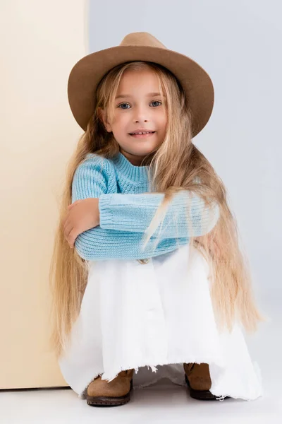 Menina loira na moda em chapéu marrom e botas, saia branca e suéter azul sentado perto da parede bege — Fotografia de Stock