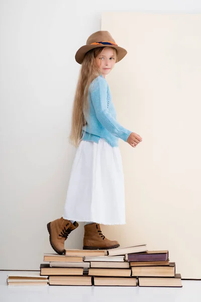Side view of fashionable blonde girl in brown hat and boots, white skirt and blue sweater walking on vintage books — Stock Photo