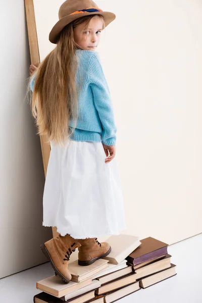 Back view of fashionable blonde girl in brown hat and boots, white skirt and blue sweater walking on vintage books — Stock Photo