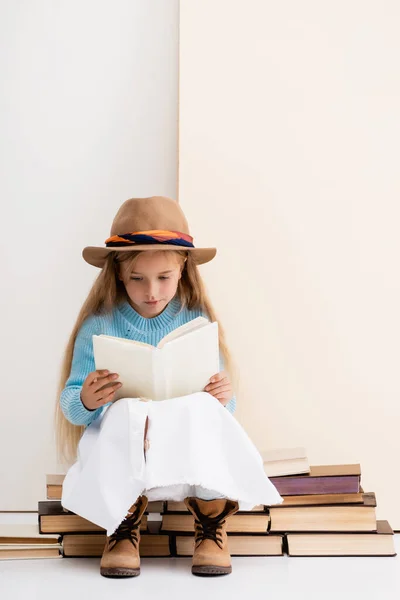 Fille blonde à la mode en chapeau et bottes marron, jupe blanche et pull bleu assis sur des livres vintage et la lecture près du mur beige — Photo de stock