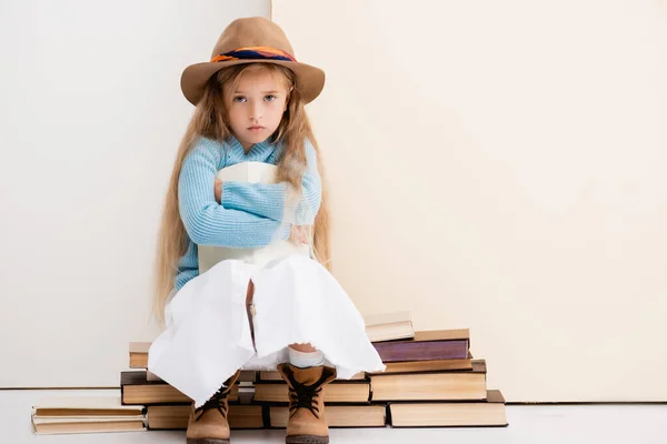 Triste fille blonde à la mode en chapeau brun et bottes, jupe blanche et pull bleu assis avec livre près du mur beige — Photo de stock