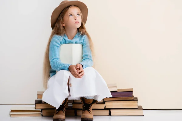Мрійлива модна блондинка в коричневому капелюсі і чоботях, біла спідниця і синій светр сидить з книгою біля бежевої стіни — стокове фото