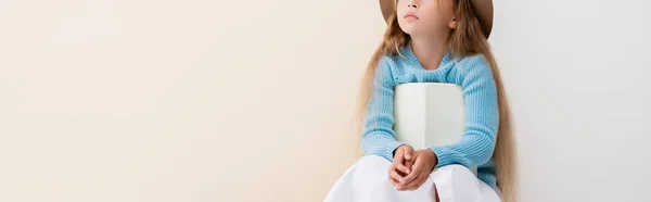 Vista recortada de la chica rubia de moda sentado con libro sobre fondo beige y blanco, plano panorámico - foto de stock