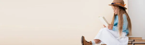 Chica rubia de moda en sombrero marrón y botas, falda blanca y suéter azul sentado en libros antiguos y lectura cerca de la pared beige, tiro panorámico - foto de stock
