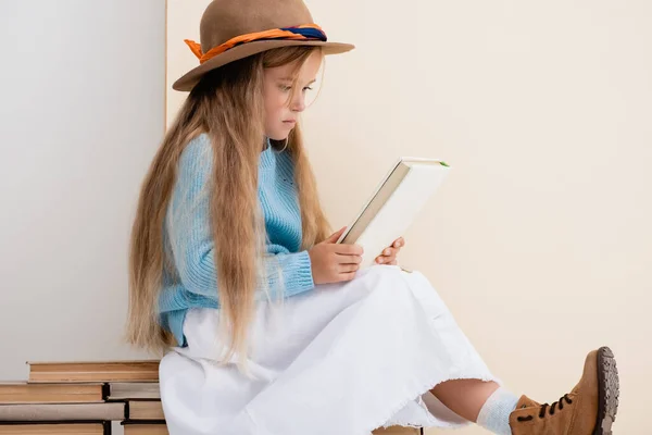 Chica rubia de moda en sombrero marrón y botas, falda blanca y suéter azul sentado en libros antiguos y leyendo cerca de la pared beige - foto de stock