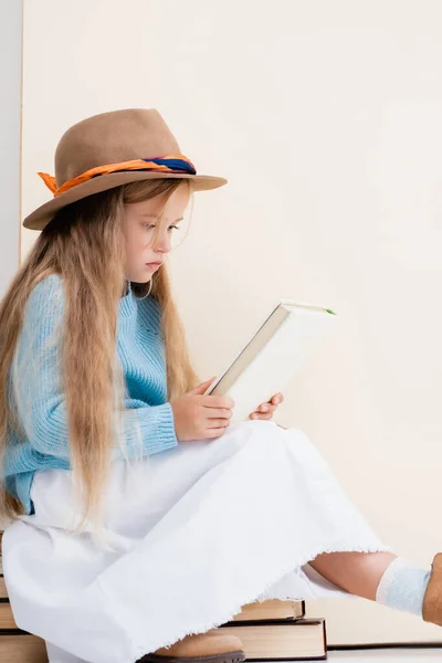 Menina loira na moda em chapéu marrom, saia branca e suéter azul sentado em livros vintage e leitura perto da parede bege — Fotografia de Stock