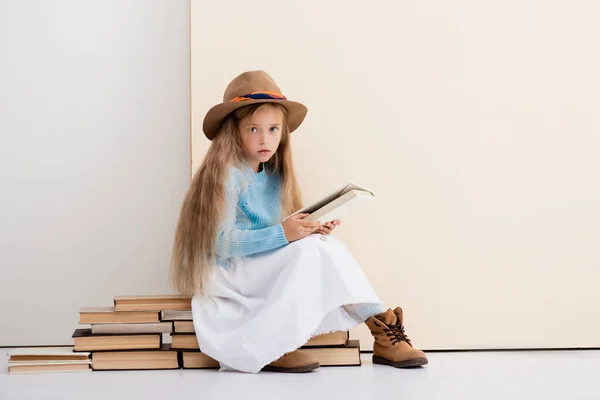 Modische blonde Mädchen mit braunem Hut und Stiefeln, weißem Rock und blauem Pullover sitzen auf Vintage-Bücher und lesen in der Nähe beige Wand — Stockfoto