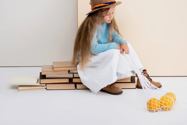 Fille blonde à la mode en chapeau brun et bottes, jupe blanche et pull bleu assis sur des livres vintage avec des pamplemousses dans un sac à ficelle — Photo de stock