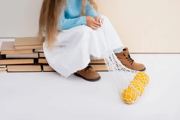 Abgeschnittene Ansicht eines modischen blonden Mädchens in braunen Stiefeln, weißem Rock und blauem Pullover, das auf Vintage-Büchern mit Grapefruits im String-Bag sitzt — Stockfoto