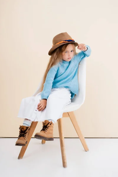 Chica rubia de moda en sombrero marrón y botas, falda blanca y suéter azul sentado en la silla cerca de la pared beige - foto de stock
