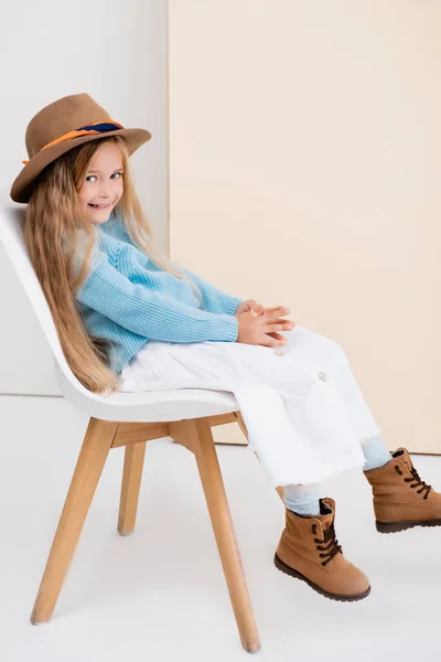 Side view of fashionable blonde girl in brown hat and boots, white skirt and blue sweater sitting on chair and smiling near beige wall — Stock Photo